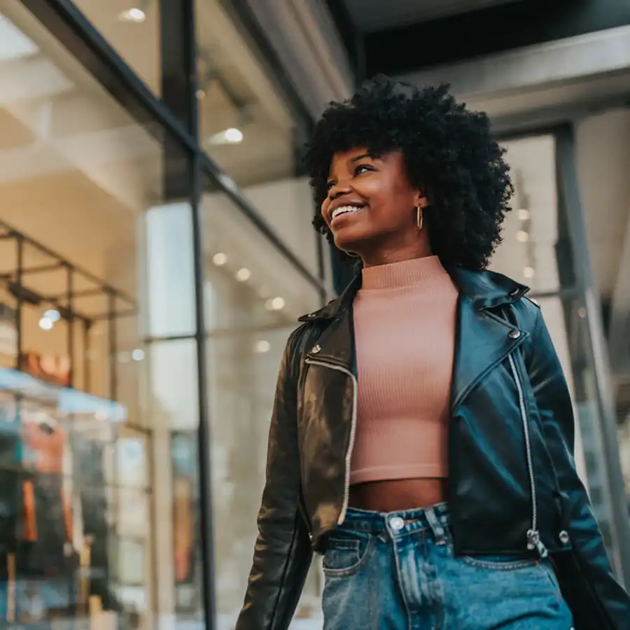 A woman walking down the street, smiling and glancing at a shop window.